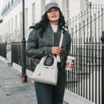 Load image into Gallery viewer, Woman walking on a sidewalk holding a cream handbag and a drink, wearing a grey jacket and black jeans.