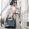 Load image into Gallery viewer, Woman in a trench coat holding a blue handbag on a city street.