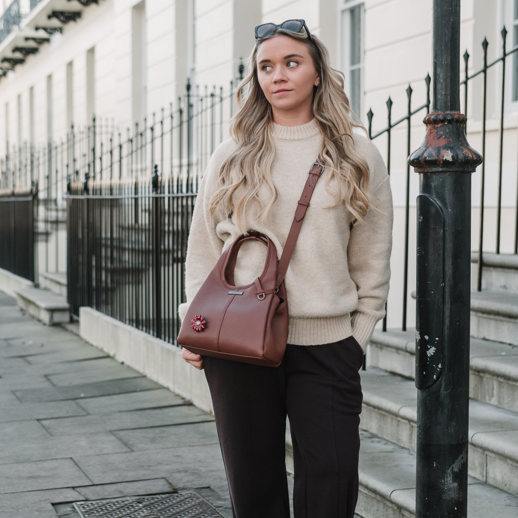 Woman in a beige sweater and dark pants holding a brown handbag on a city street.