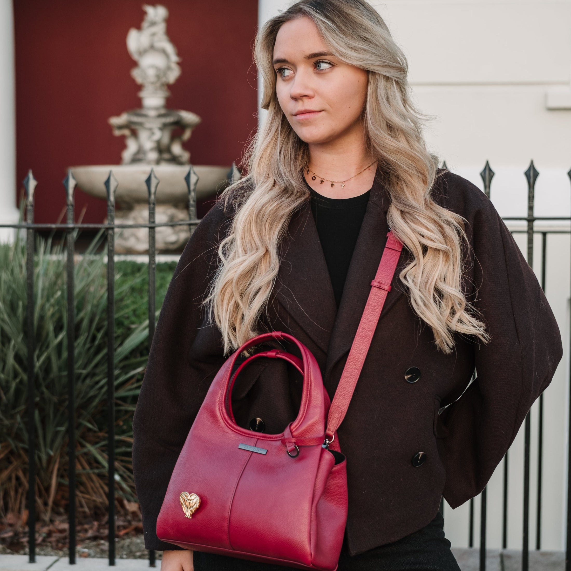 Woman holding a red handbag in front of a building with a fountain.